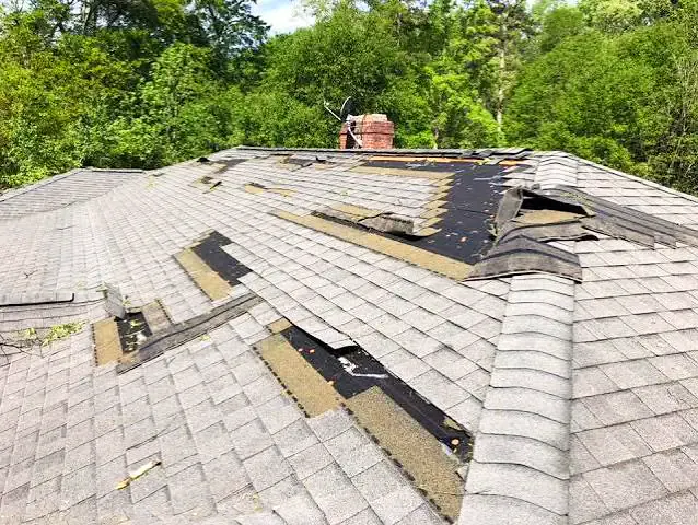 Asphalt shingles lifted, creased and torn away from a roof slope around a brick chimney after high winds